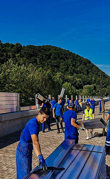 Group of workers in blue uniforms installing large metal panels along a roadside barrier. The scene includes a paved walkway, vehicles on the adjacent road, and green hills in the background under a clear blue sky.
