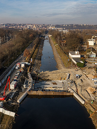 Aerial view of a river navigation channel under construction, featuring a partially built lock structure with steel sheet piling, heavy machinery, and construction materials along the banks. The surrounding area includes a parallel railway line, access roads, and scattered buildings, with a wide river visible to the right and a distant cityscape on the horizon.