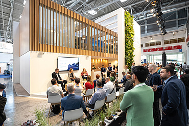Visitors attending a panel discussion at the STRABAG and WTE exhibition stand at IFAT, featuring a modern booth design with wood elements, greenery, and a presentation area.