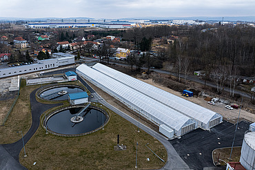 Luftaufnahme einer Kläranlage mit zwei runden Klärbecken und langen, gewächshausähnlichen Strukturen, die zur solaren Schlammtrocknung genutzt werden, umgeben von Straßen, Grünflächen und nahe gelegenen Wohn- und Industriegebäuden.