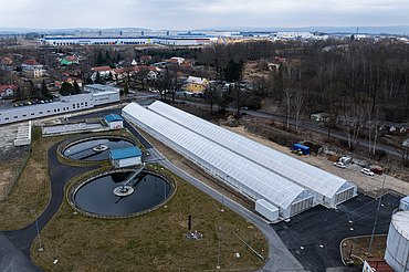 Luftaufnahme einer Kläranlage mit zwei runden Klärbecken und langen, gewächshausähnlichen Strukturen, die zur solaren Schlammtrocknung genutzt werden, umgeben von Straßen, Grünflächen und nahe gelegenen Wohn- und Industriegebäuden.