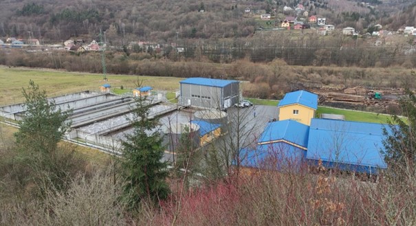 Luftaufnahme einer Wasseraufbereitungsanlage mit Gebäuden mit blauen Dächern und rechteckigen Becken, umgeben von Bäumen und Hügeln in einer ländlichen Gegend.
