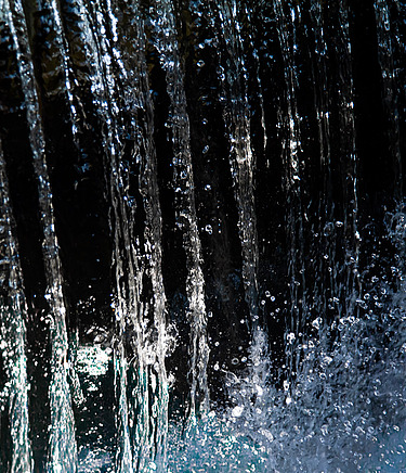 Close-up view of cascading water streams falling vertically, with droplets splashing and shimmering in the light against a dark background.