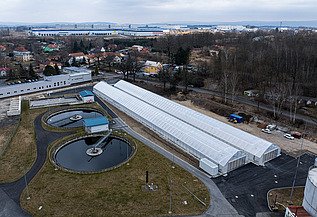Luftaufnahme einer Kläranlage mit zwei runden Klärbecken und langen, gewächshausähnlichen Strukturen, die zur solaren Schlammtrocknung genutzt werden, umgeben von Straßen, Grünflächen und nahe gelegenen Wohn- und Industriegebäuden.