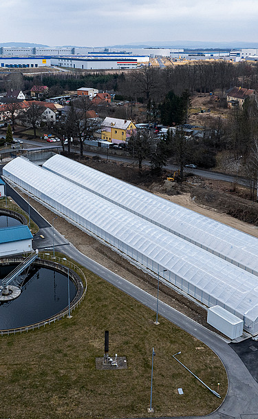 Luftaufnahme einer Kläranlage mit zwei runden Klärbecken und langen, gewächshausähnlichen Strukturen, die zur solaren Schlammtrocknung genutzt werden, umgeben von Straßen, Grünflächen und nahe gelegenen Wohn- und Industriegebäuden.