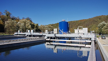 Mit Wasser gefülltes Betonbecken in einer Kläranlage mit Metallstegen, Rohren und einem großen blauen zylindrischen Tank, umgeben von Bäumen und Hügeln unter einem klaren Himmel.