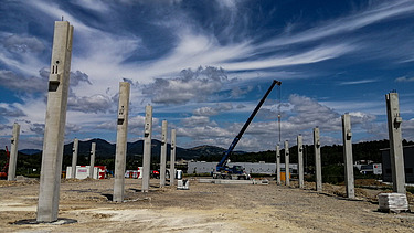 Construction site with multiple upright concrete columns arranged in rows, forming the framework of a large structure. A crane is positioned in the center, lifting materials. The background shows hills, scattered buildings, and a partly cloudy sky with dramatic cloud patterns.