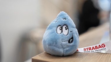Close-up of a friendly, blue water droplet plush toy with a STRABAG ribbon, serving as a charming giveaway.