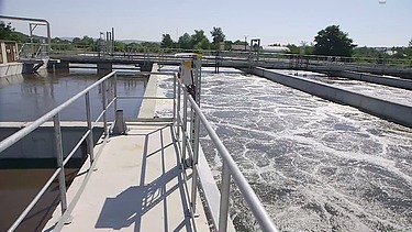 View along a walkway at a wastewater treatment plant showing aeration basins with bubbling water, metal railings, pipes, and treatment equipment under clear daylight.
