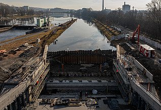 Construction site of a navigation channel lock along a river, showing reinforced concrete structures, steel sheet piling, and workers, with waterway extending into the background and urban buildings visible on the horizon.