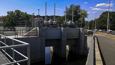Concrete floodgate structure with three openings and metal railings, located next to a road. The structure includes vertical control rods and mechanical equipment on top. Trees and streetlights line the background under a clear blue sky.