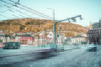 Cobblestone city square with tram tracks, colorful historic buildings, and motion-blurred vehicles, set against green hills.
