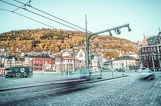 Gepflasterter Stadtplatz mit Straßenbahnschienen, farbenfrohen historischen Gebäuden und Fahrzeugen mit Bewegungsunschärfe vor grüner Hügellandschaft.