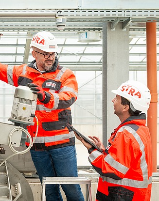 Zwei Arbeiter in orange-schwarzen Sicherheitsjacken und weißen Schutzhelmen in einer Industrieanlage, die ein mechanisches Bauteil überprüfen und bedienen, wobei eine Person ein Tablet hält und die andere einen Motor einstellt.