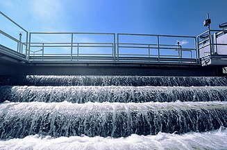 Mehrstufige Wasserkaskade in einer Kläranlage mit schäumendem Wasser, das über Stufen unter einer Metallgeländerkonstruktion unter blauem Himmel fließt.