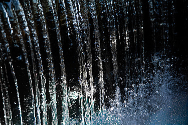 Close-up view of cascading water streams falling vertically, with droplets splashing and shimmering in the light against a dark background.