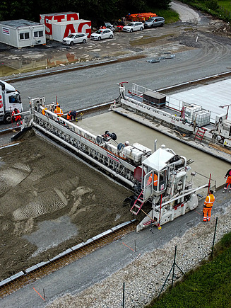 Baustelle einer Kläranlage mit Arbeitern und schweren Maschinen, die große Betonplatten für solare Klärschlammtrocknungsanlagen installieren, mit Lastwagen und Geräten im Hintergrund.