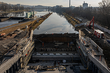 Baustelle einer Fahrrinnenschleuse an einem Fluss mit Stahlbetonkonstruktionen, Stahlspundwänden und Arbeitern. Im Hintergrund ist die Wasserstraße zu sehen, am Horizont sind städtische Gebäude zu erkennen.