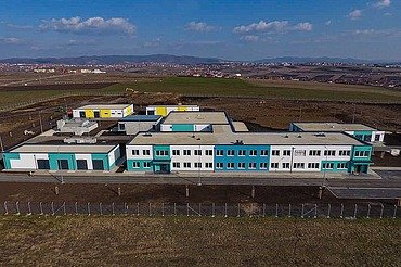 Luftaufnahme einer modernen Wasseraufbereitungsanlage in Prishtina mit mehreren Flachbauten mit weißen und türkisfarbenen Fassaden, Flachdächern und umliegenden gepflasterten Straßen. Die Anlage befindet sich auf einer großen Freifläche, auf der im Hintergrund Baugeräte zu sehen sind, sowie in der Ferne Hügel unter einem teilweise bewölkten Himmel.