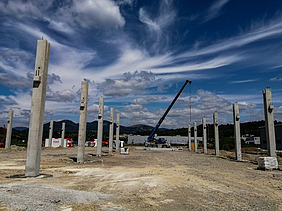 Baustelle mit mehreren aufrecht stehenden Betonsäulen, die in Reihen angeordnet sind und das Gerüst eines großen Bauwerks bilden. In der Mitte steht ein Kran, der Materialien anhebt. Der Hintergrund zeigt Hügel, verstreute Gebäude und einen teilweise bewölkten Himmel mit dramatischen Wolkenmustern.