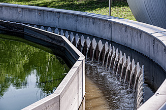 Circular clarifier basin at a wastewater treatment plant with overflow weirs; clear water flows over the serrated edge while adjacent basin contains green water – photographed at WWTP Korneuburg.