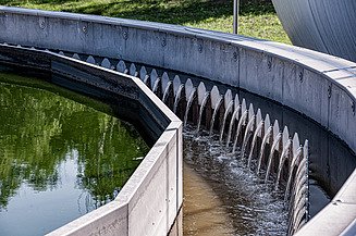 Circular clarifier basin at a wastewater treatment plant with overflow weirs; clear water flows over the serrated edge while adjacent basin contains green water – photographed at WWTP Korneuburg.