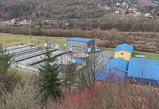 Aerial view of a water treatment facility with blue-roofed buildings and rectangular basins, surrounded by trees and hills in a rural area.