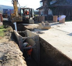 Bagger und Arbeiter beim Verlegen einer Pipeline in einem tiefen Graben mit Schutzverbau entlang einer Wohnstraße.