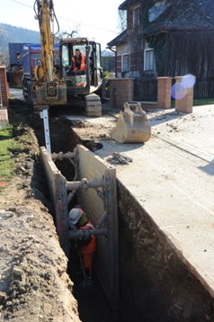 Bagger und Arbeiter beim Verlegen einer Pipeline in einem tiefen Graben mit Schutzverbau entlang einer Wohnstraße.