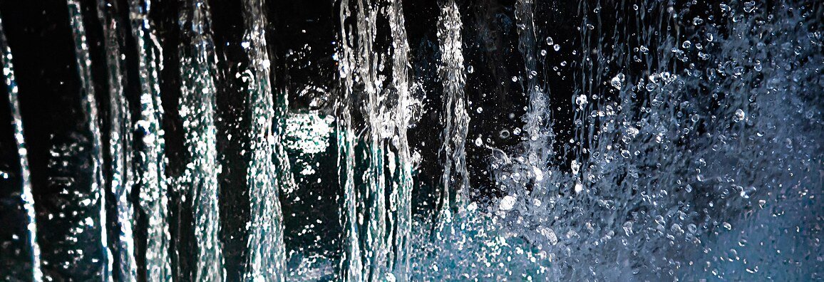Close-up view of cascading water streams falling vertically, with droplets splashing and shimmering in the light against a dark background.
