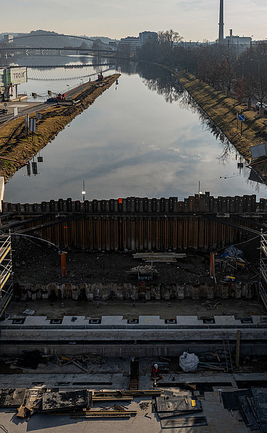 Baustelle einer Fahrrinnenschleuse an einem Fluss mit Stahlbetonkonstruktionen, Stahlspundwänden und Arbeitern. Im Hintergrund ist die Wasserstraße zu sehen, am Horizont sind städtische Gebäude zu erkennen.