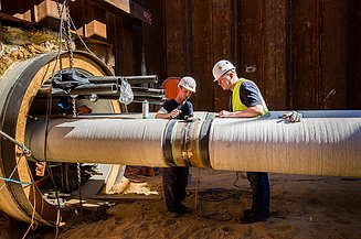 Two workers inspecting and assembling a large insulated pipeline at an industrial construction site.