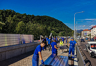 Gruppe von Arbeitern in blauen Uniformen, die große Metallplatten entlang einer Straßenabsperrung montieren. Die Szene umfasst einen gepflasterten Gehweg, Fahrzeuge auf der angrenzenden Straße und grüne Hügel im Hintergrund unter einem klaren blauen Himmel.