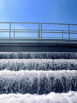 Multi-tiered water cascade at a treatment facility, with foaming water flowing over steps beneath a metal railing structure under a clear blue sky.