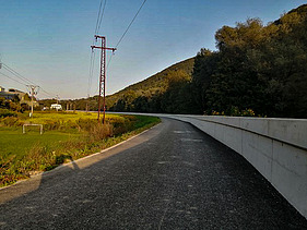 Gerade asphaltierte Straße, die an einer hohen Hochwasserschutzmauer aus Beton entlangführt. Auf der linken Seite befindet sich ein grasbewachsenes Gelände mit Stromleitungen und -masten sowie ein paar Häusern in der Ferne. Auf der rechten Seite erhebt sich ein bewaldeter Hügel unter klarem Himmel.