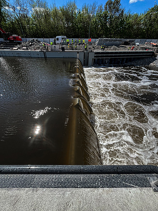 Close-up view of a river lock under construction, showing water flowing over a series of curved barriers creating a controlled cascade, with turbulent water on the right side. In the background, workers in safety gear and construction equipment are visible along the concrete structure, surrounded by trees under a clear blue sky.