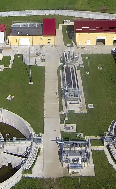 Aerial view of the Moinești wastewater treatment plant in Romania, showing circular clarifiers, treatment basins, control buildings with red roofs, and surrounding green fields within a fenced site.