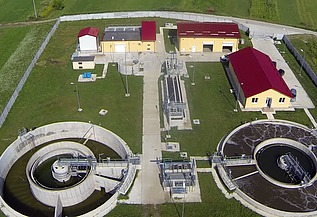Aerial view of the Moinești wastewater treatment plant in Romania, showing circular clarifiers, treatment basins, control buildings with red roofs, and surrounding green fields within a fenced site.