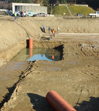 Baustelle mit Arbeitern, die große orangefarbene Drainagerohre verlegen und eine rechteckige, mit Wasser gefüllte Baugrube.