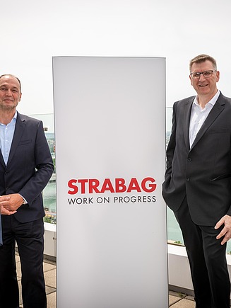 The Executive Board members of STRABAG and WTE Wassertechnik posing for a group photo at Donau-City-Straße. They are standing next to a STRABAG brand sign, symbolizing the integration of WTE into the UB4K Sub Division LC Water.