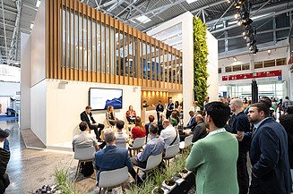 Besucher einer Podiumsdiskussion auf dem Messestand von STRABAG und WTE auf der IFAT, der sich durch ein modernes Standdesign mit Holzelementen, Begrünung und einem Präsentationsbereich auszeichnet.