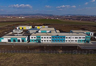 Aerial view of a modern water treatment plant in Prishtina, featuring multiple low-rise buildings with white and turquoise facades, flat roofs, and surrounding paved roads. The facility is located in a large open area with construction equipment visible in the background and distant hills under a partly cloudy sky.