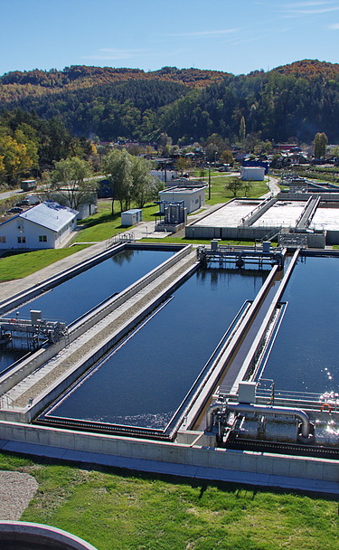Aerial view of a wastewater treatment plant with multiple large rectangular basins, surrounded by green grass, trees, and rolling hills.