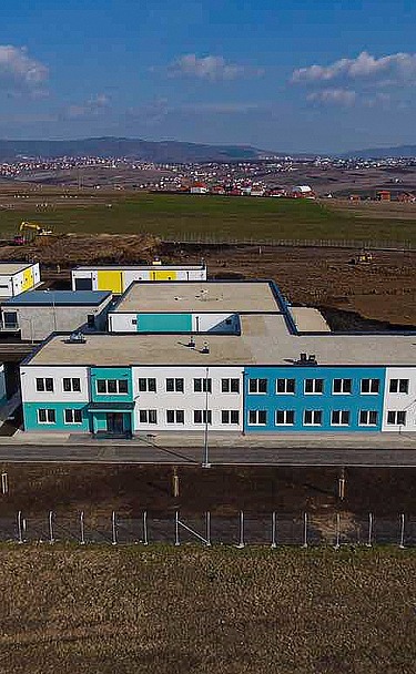 Luftaufnahme einer modernen Wasseraufbereitungsanlage in Prishtina mit mehreren Flachbauten mit weißen und türkisfarbenen Fassaden, Flachdächern und umliegenden gepflasterten Straßen. Die Anlage befindet sich auf einer großen Freifläche, auf der im Hintergrund Baugeräte zu sehen sind, sowie in der Ferne Hügel unter einem teilweise bewölkten Himmel.