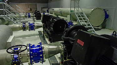 Innenansicht einer Industrieanlage mit großen Rohrleitungen, blauen Absperrventilen und mehreren schwarzen Pumpenmotoren, die auf einem Betonboden montiert sind. Im Hintergrund sind weitere Rohre und eine Metalltreppe zu sehen.