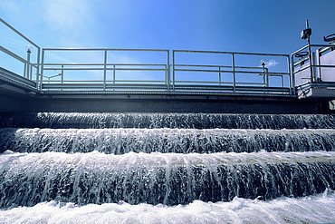 Mehrstufige Wasserkaskade in einer Kläranlage mit schäumendem Wasser, das über Stufen unter einer Metallgeländerkonstruktion unter blauem Himmel fließt.