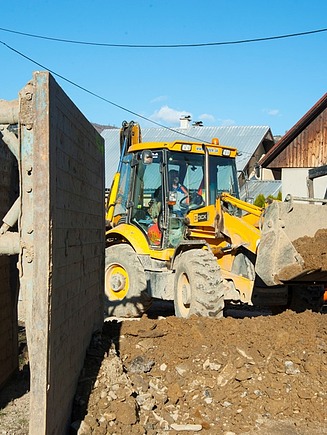 Gelber Baggerlader, der auf einer Baustelle mit Grabenverbauplatten und orangefarbenen Entwässerungsrohren in der Nähe von Wohnhäusern Erde bewegt.