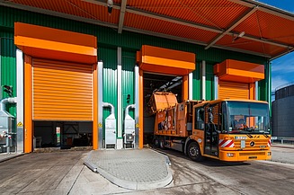 Ein orangefarbener Müllwagen ist in einem modernen Recyclingzentrum mit grünen und orangefarbenen Industriehallen zu sehen. Das Fahrzeug steht an einer offenen Laderampe für die Abfallentsorgung.