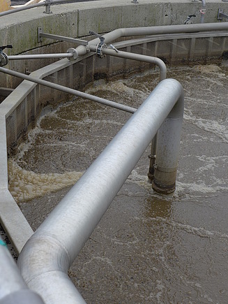 Circular wastewater treatment tank with turbulent brown water and integrated stainless steel piping system. The pipes are connected to the tank wall and extend across the structure for aeration or water distribution.