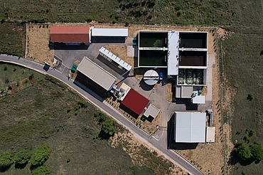 Draufsicht auf eine Kläranlage mit mehreren rechteckigen Klärbecken, einem runden Klärbecken und mehreren Gebäuden mit roten und weißen Dächern, umgeben von grünen Feldern und Zufahrtsstraßen.